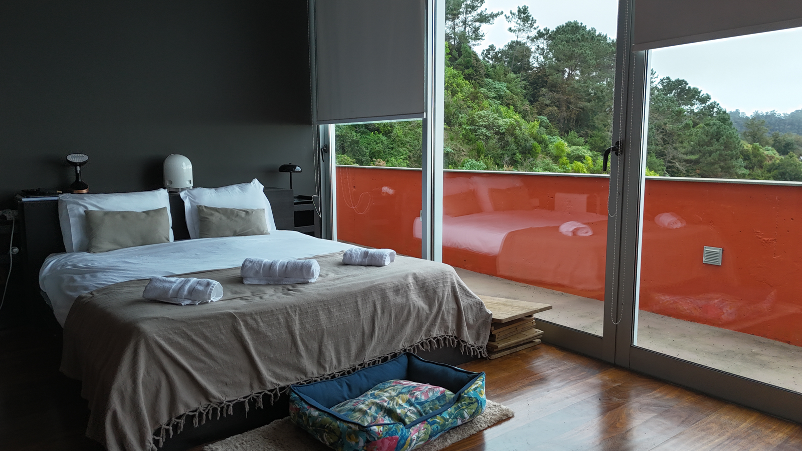 Master bedroom with a charcoal wall, white bed, orange balcony, and forest view
