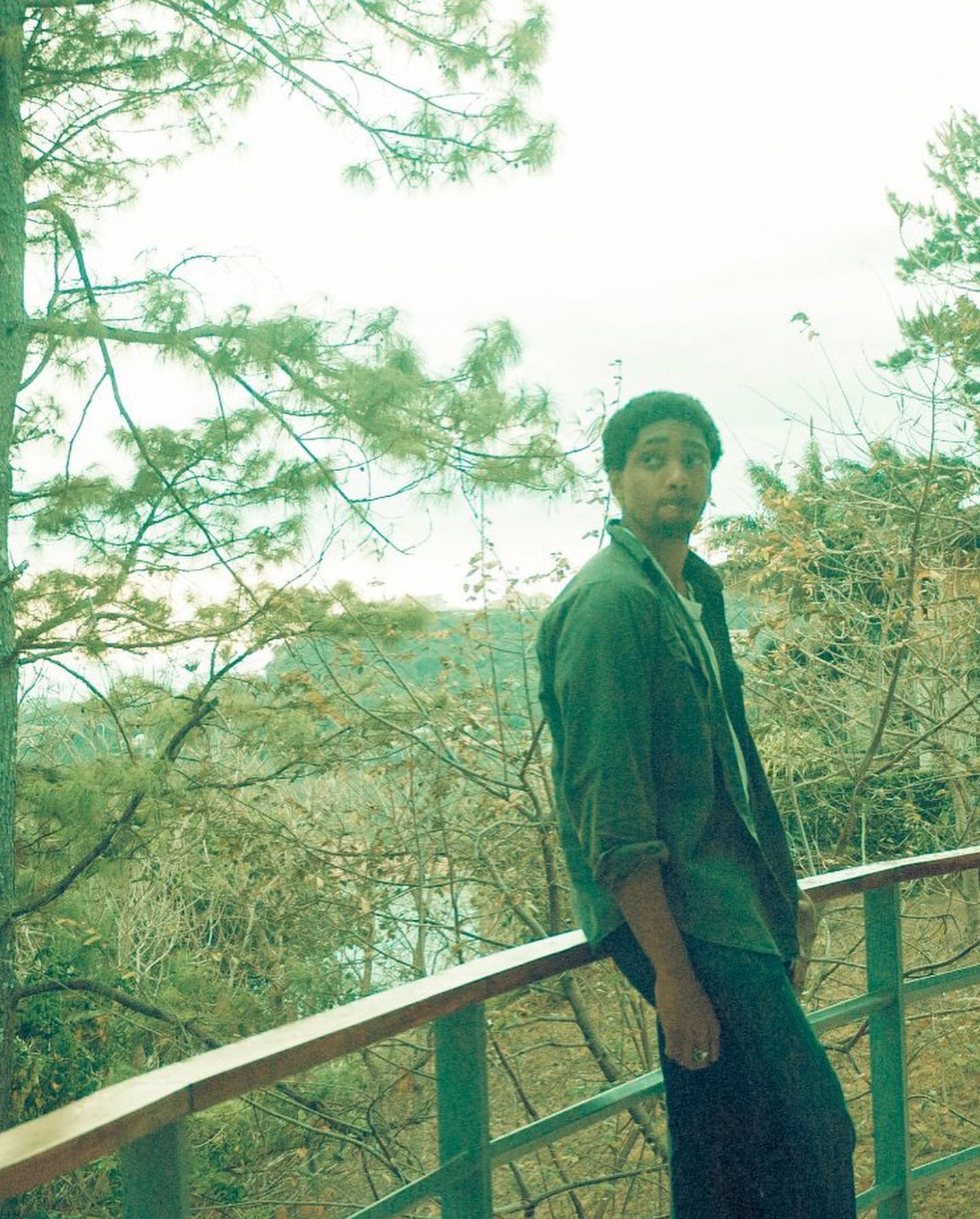 Keem leaning on a balcony rail, pines behind him